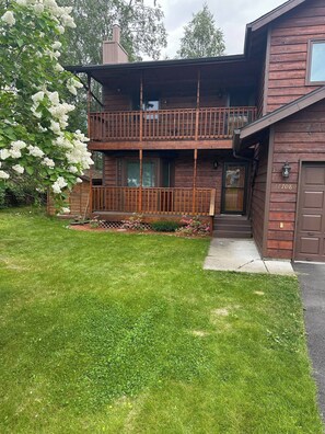 A two-story wooden house with a balcony, a well-maintained lawn, and a tree with white flowers.