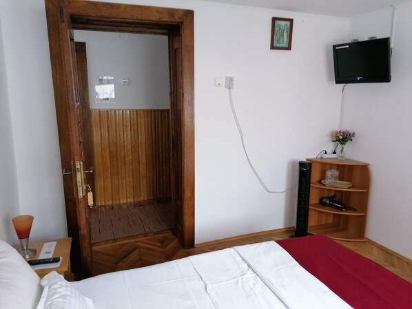 A hotel room with a wooden door, a bed with white and red bedding, a TV mounted on the wall, and a small wooden shelf with a vase and a hairdryer. - Pensiunea Ioana (Curtea De Arges)