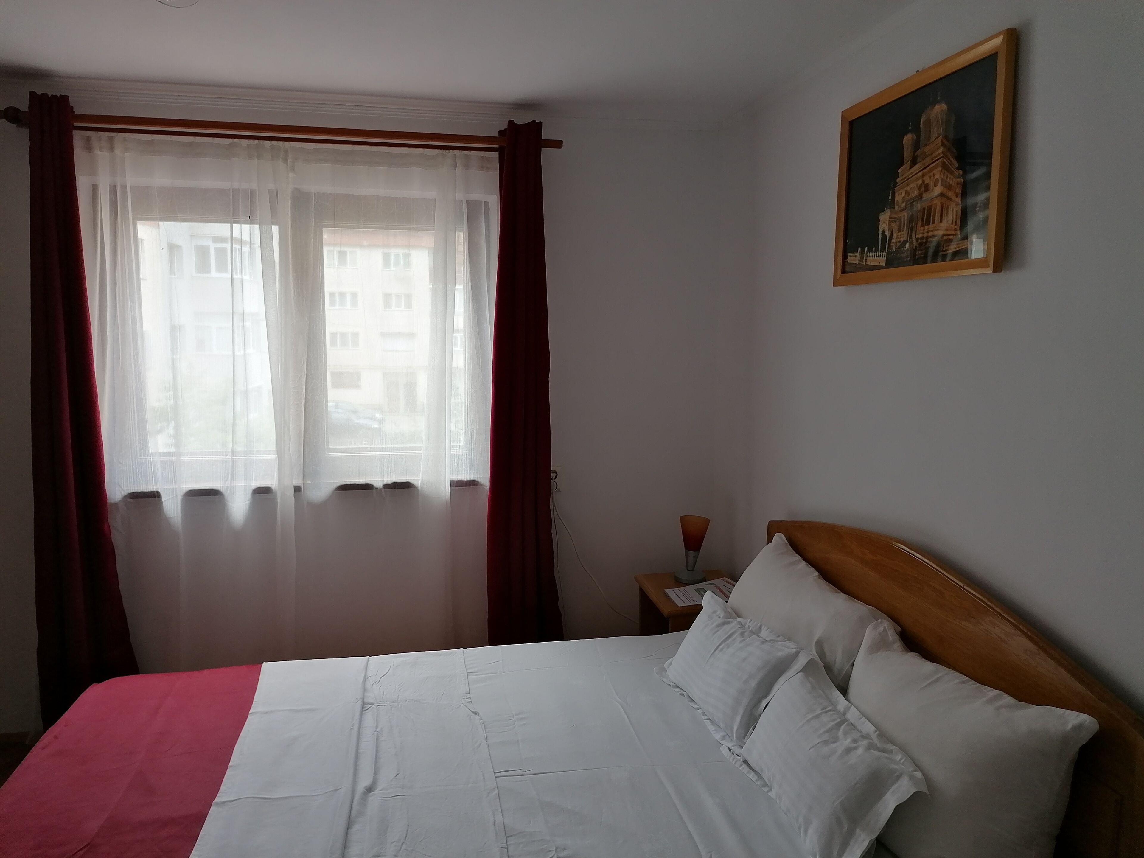 Una habitación de hotel con cama, mesita de noche, un cuadro en la pared y una ventana con cortinas traslúcidas.