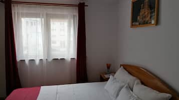 Una habitación de hotel con cama, mesita de noche, un cuadro en la pared y una ventana con cortinas traslúcidas.