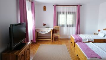 A room with a television on a wooden cabinet, a desk with a chair, a bed with a purple blanket, and a window with curtains.