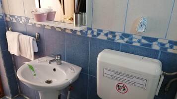 A bathroom with a sink, mirror, and toilet. There is a towel rack with a white towel, a soap dispenser, and a trash bin. The walls are tiled with blue and white patterns.