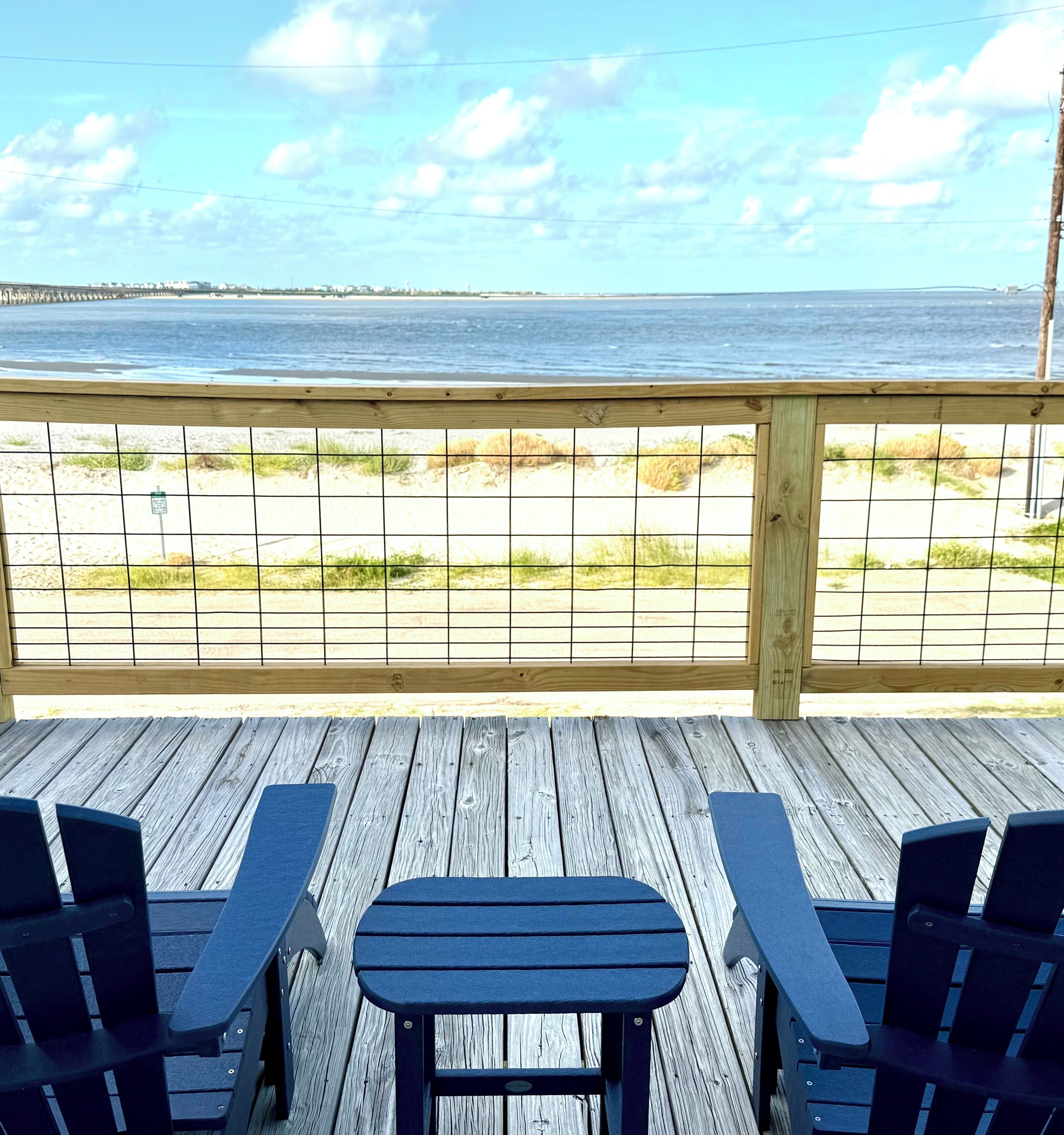A wooden deck with two tables and chairs, overlooking a beach with a wooden pier in the distance.