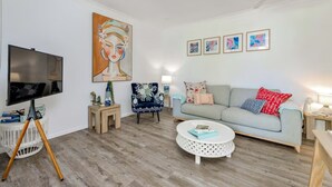 Una sala de estar con una televisión de pantalla plana, un sillón azul, un sofá gris claro y una mesa de centro redonda.
