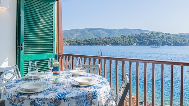 A balcony with a table set for dining, overlooking a body of water with boats and hills in the background. - Sea view apartment in Porto Azzurro (Porto Azzurro)