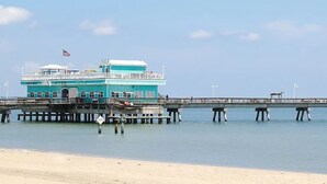 A turquoise building with a balcony, a pier extending into the water, and a sandy beach in the foreground. - Sojourn The Lighthouse  Beach Front Resort Villa (Norfolk)