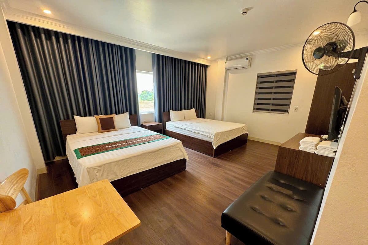 Ein Hotelzimmer mit zwei Betten, einem Holztisch, einer Bank, einem Ventilator und einem Fenster mit Vorhängen.
