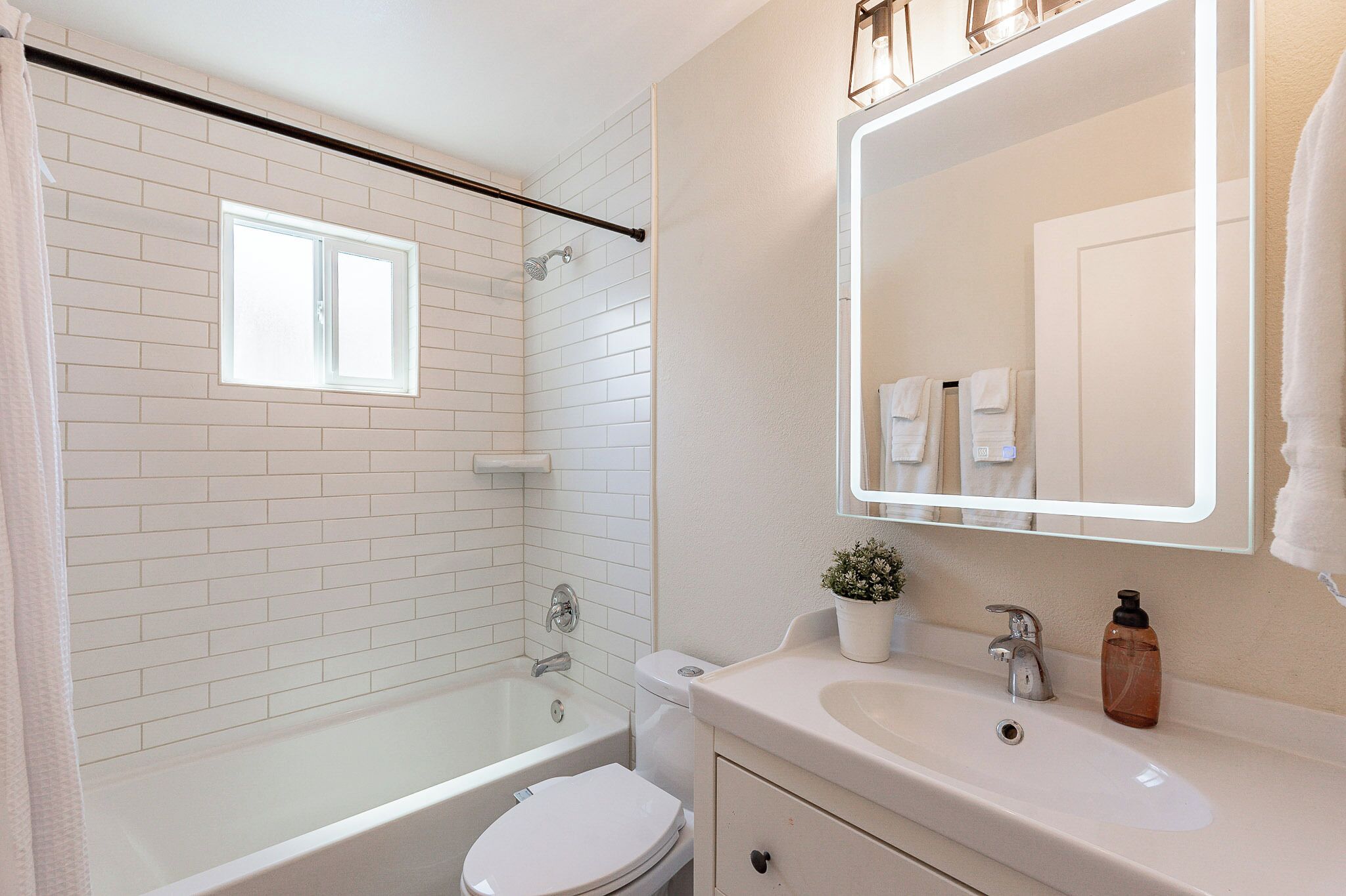 A modern bathroom with a white bathtub, toilet, and sink. There is a window above the bathtub and a mirror above the sink.