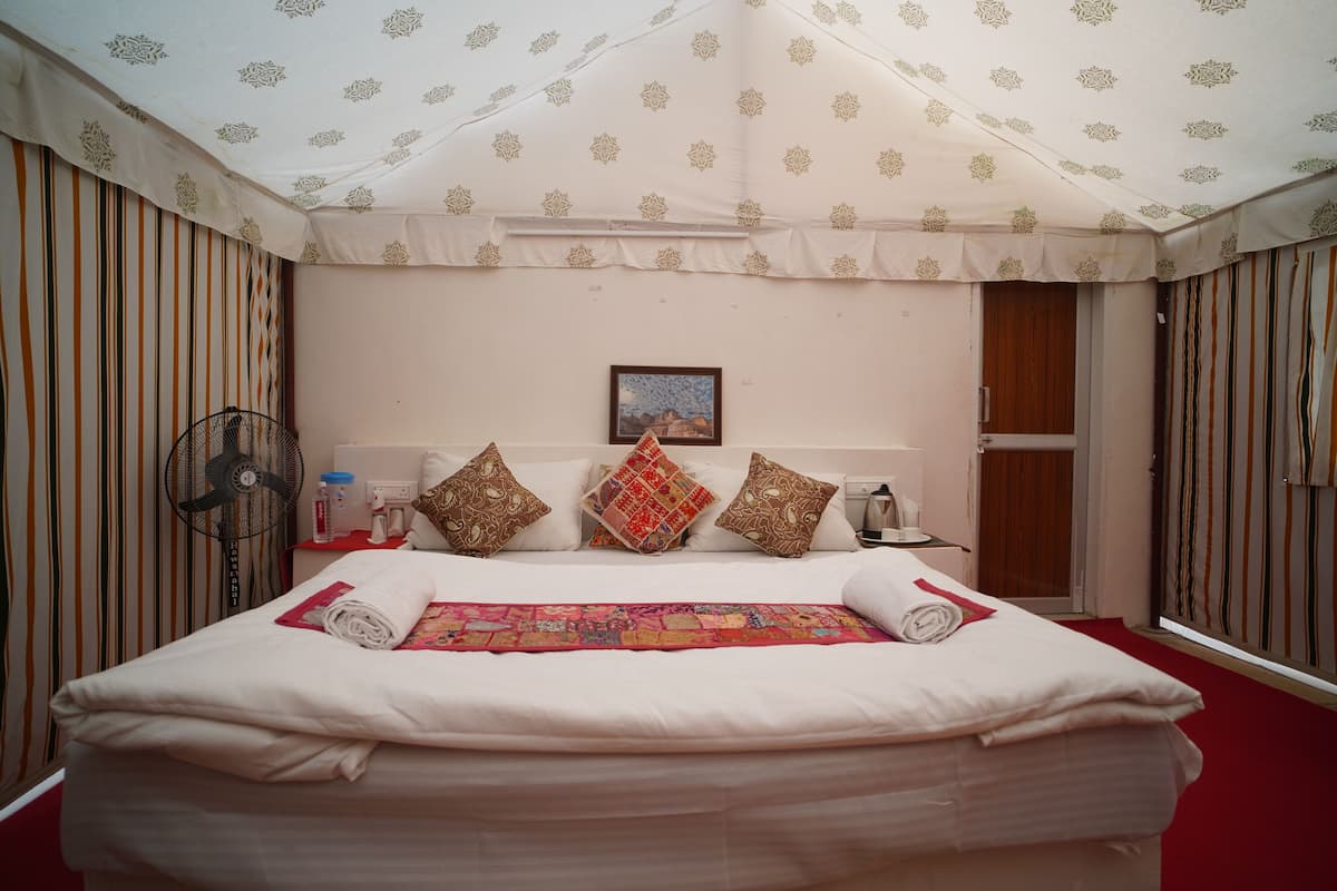A neatly made bed with a colorful blanket and pillows, a bedside table with a fan and a bottle, and a framed picture on the wall.