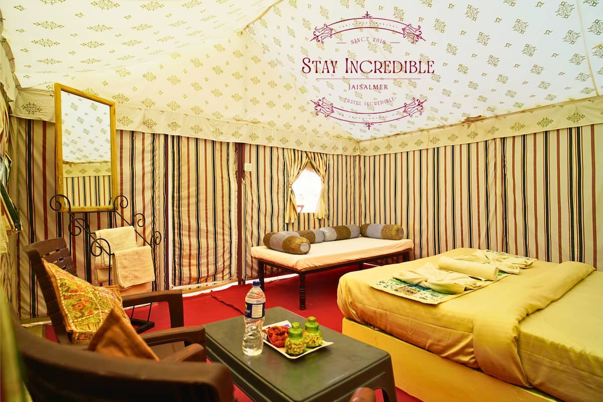 A hotel room with striped curtains, a bed with a yellow bedspread, a bedside table with a bottle and snacks, a chair with a cushion, and a mirror on the wall.