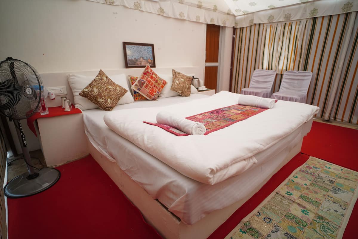 A neatly made bed with a red and white blanket, a standing fan, and a patterned rug.