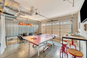 A ping pong table with stools, a seating area with cushions, and a large window with curtains.