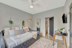 A modern bedroom with a tufted bed, a standing lamp, a flat-screen TV, and a small table with plants.