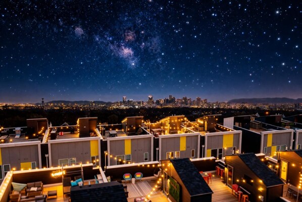 A rooftop terrace with string lights, a city skyline in the background, and a starry night sky.