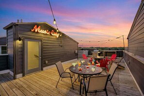 A rooftop patio with a neon sign reading "Music City," a round table with drinks, and red chairs.