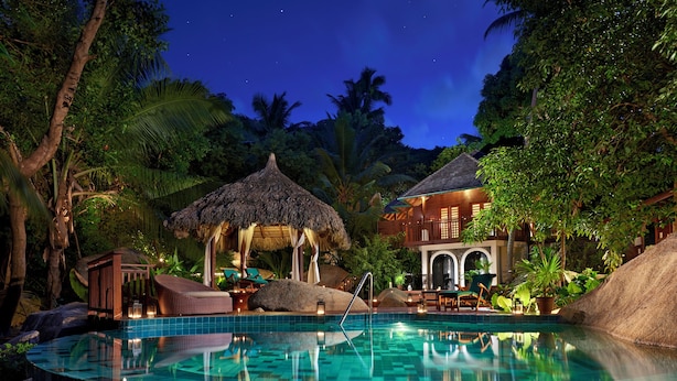 A nighttime pool area with a thatched-roof structure, lounge chairs, and a building with arched windows. - Niva Labriz Seychelles (Silhouette Island)