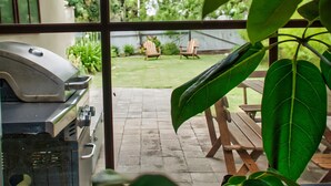 Un espace patio aménagé avec un barbecue, des meubles en bois et une vue sur un jardin avec une pelouse et un fauteuil.