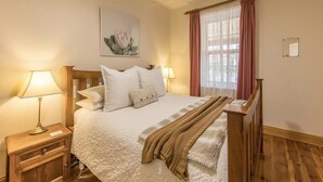 Un lit bien fait, recouvert d’une couette blanche, avec deux lampes de chevet, une tête de lit en bois, une fenêtre ornée de rideaux roses et une œuvre d’art encadrée accrochée au mur.
