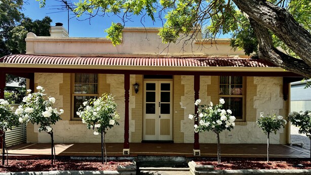 Un bâtiment à deux étages, avec un toit brun, des murs de couleur beige et un porche à l’avant orné de plantes à fleurs blanches.