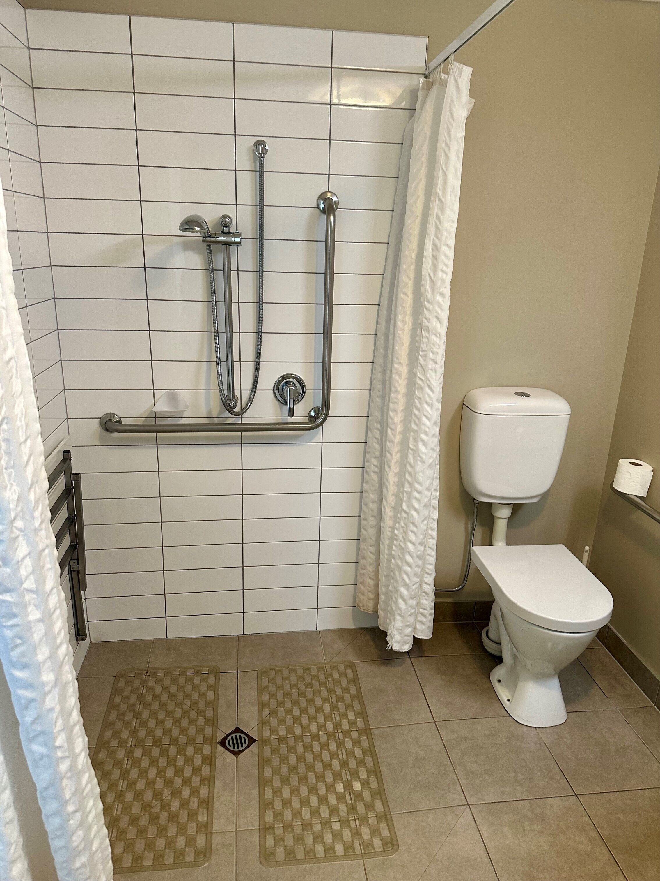 A bathroom with white tiled walls, a white toilet, a shower with grab bars, and a white curtain.