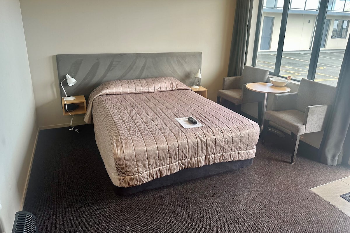 A hotel room with a large bed, a bedside table with a lamp, a chair, a small table with a bowl, and a view of the parking lot through the window.
