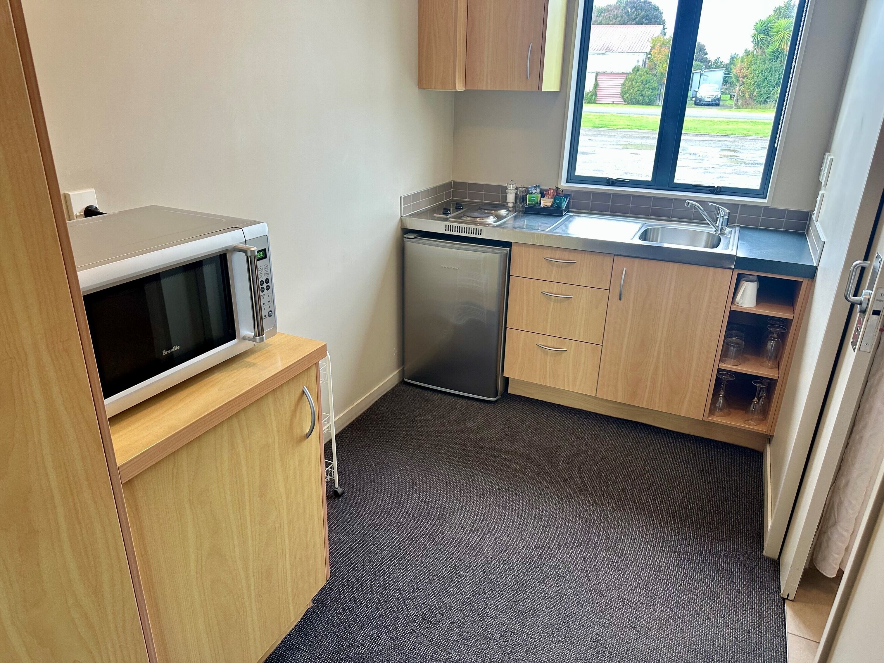 Een compacte keuken met een Breville magnetron, een RVS vaatwasser, houten kastjes en een raam met uitzicht.