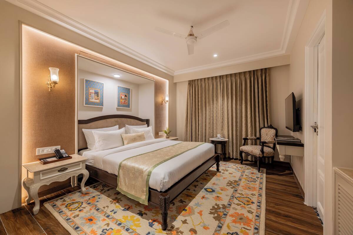 A hotel room with a large bed, a bedside table with a phone, a chair, a TV, and a decorative rug.