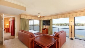 A spacious living room with a large sofa set, a wooden coffee table, and a ceiling fan. The room features large windows with a view of a body of water and a balcony. - 2BR Resort Suite in Orlando w/ Hot Tub in Suite, Resort Pools, & Near Universal (Orlando)