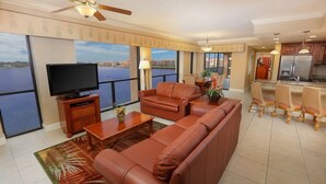 A spacious living room with a large sofa, a coffee table, and a television. There is a balcony with a view of the water and buildings. - 2BR Resort Suite in Orlando w/ Hot Tub in Suite, Resort Pools, & Near Universal (Orlando)