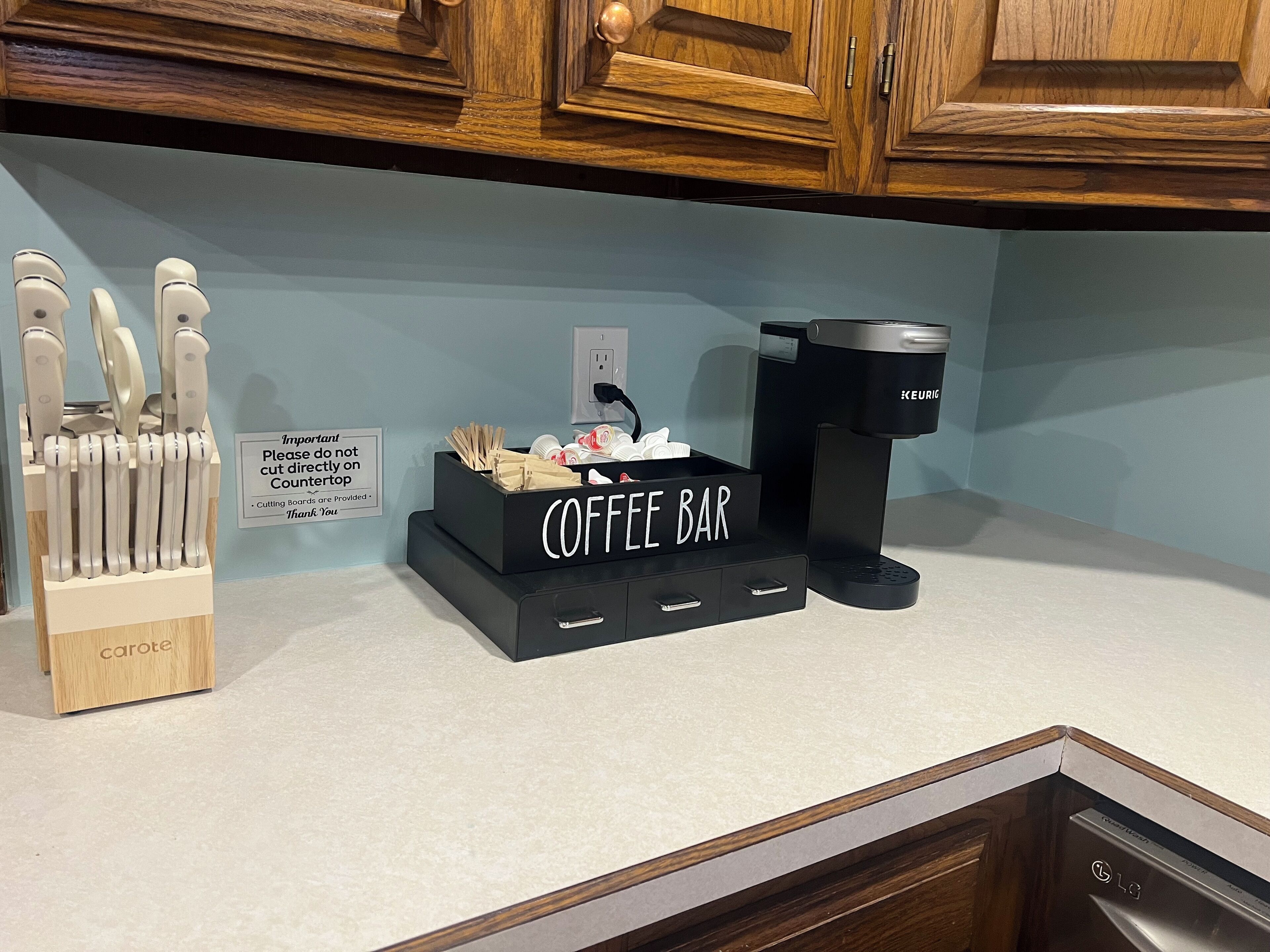 A kitchen counter with a coffee bar setup, including a coffee machine, a knife block, and a sign advising not to cut directly on the countertop.