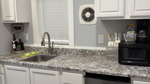 Une cuisine avec des meubles blancs, un plan de travail en granit, un lave-vaisselle en acier inoxydable, un micro-ondes noir et une fenêtre avec des stores.