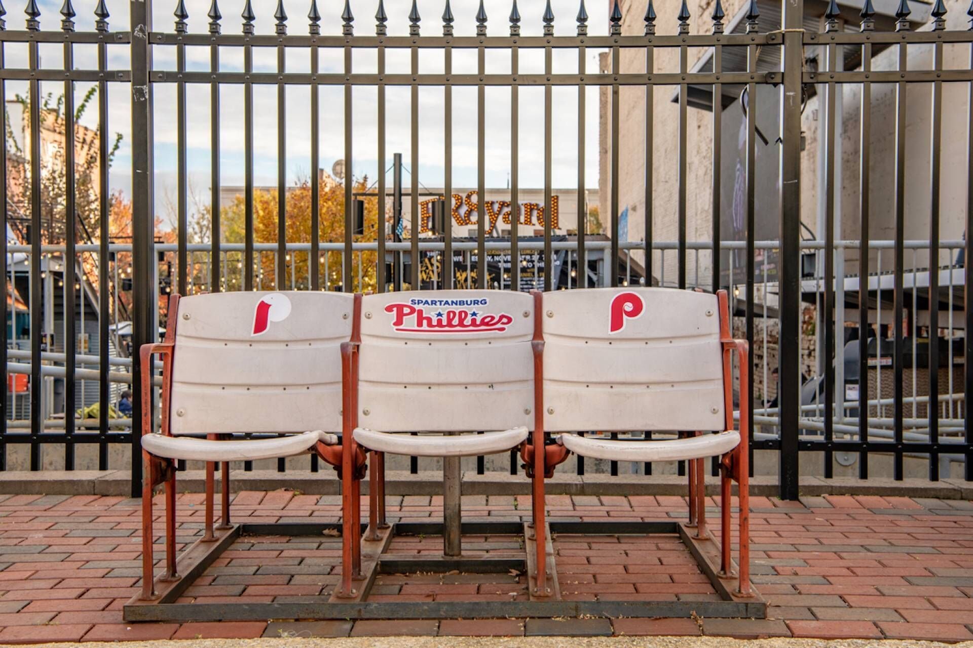 Three white park benches with red and blue Philadelphia Phillies logos, placed in front of a metal fence with a sign reading "SPARTANBURG."