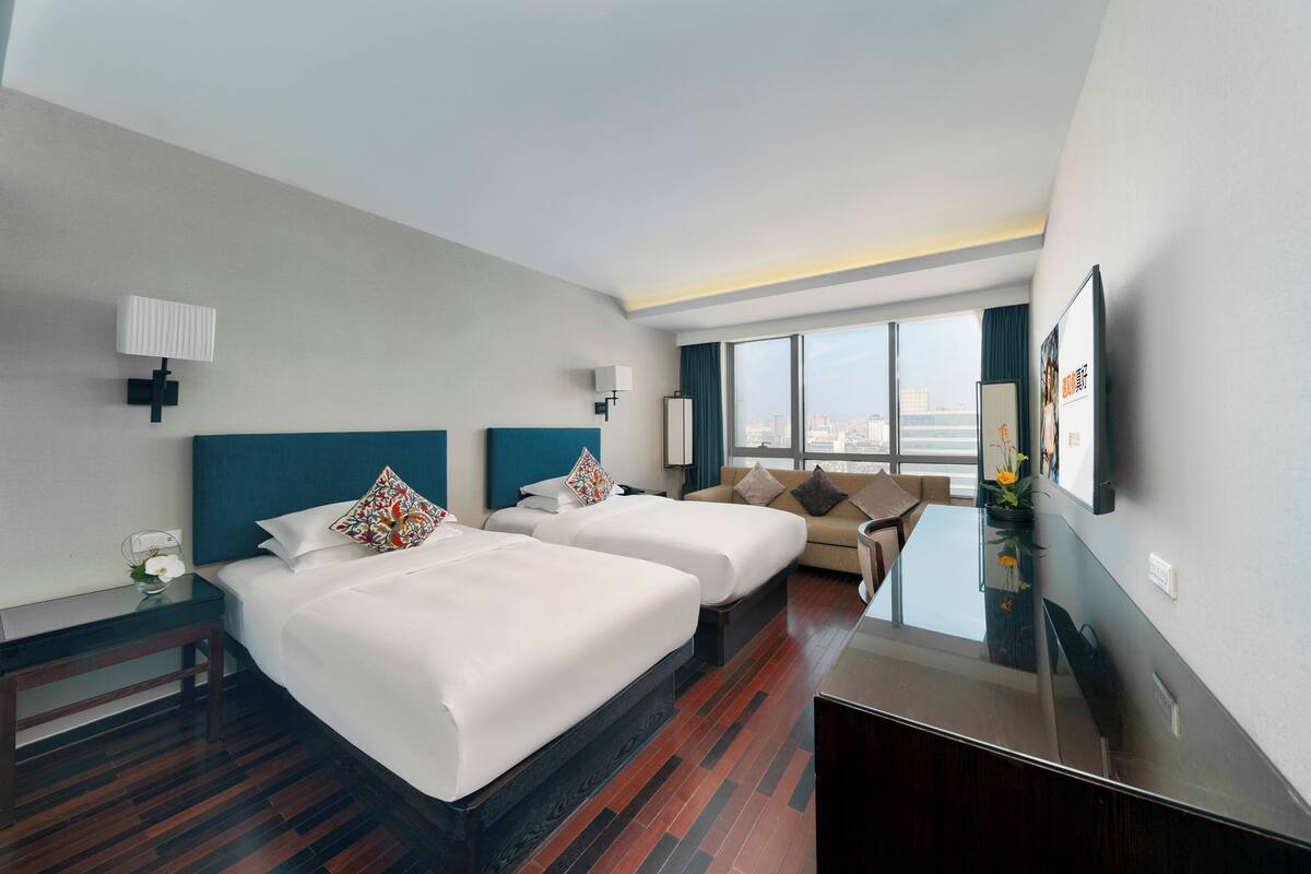 Camera d'albergo con due letti, un divano, una TV e un'ampia finestra con vista sulla città.