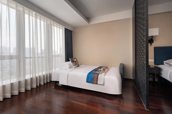 Habitación de hotel con una cama grande, un escritorio y vistas a la ciudad a través de cortinas translúcidas.