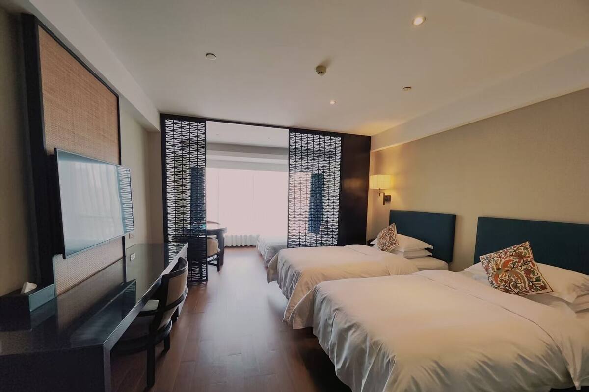 Camera d'albergo con due letti, una scrivania, una televisione e vista attraverso un pannello decorato.