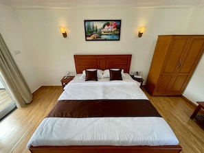 A hotel room with a large bed, wooden headboard, two bedside tables, a nightstand, a painting on the wall, and a wardrobe. - Palm Paradise Resort (Querim)