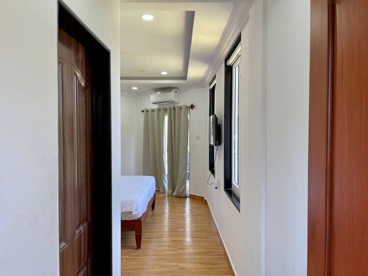 A hallway leading to a bedroom with a bed, a window with curtains, and an air conditioning unit.