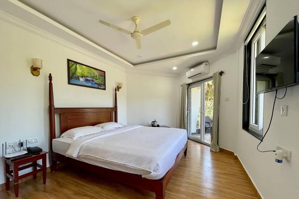 Un dormitorio con una cama de madera, una mesita de noche, un ventilador de techo, un televisor montado y una ventana con vistas.