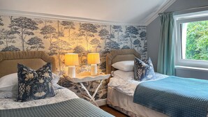 A bedroom with two beds, a nightstand with lamps, and a window with curtains. - Gillhead Farm Cottage (Matterdale End)