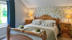 A bedroom with a wooden bed, floral wallpaper, two bedside tables with lamps, and a window with curtains. - Gillhead Farm Cottage (Matterdale End)