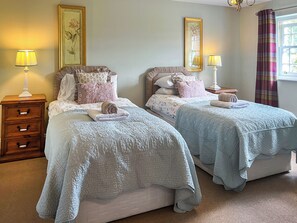 A double room with two beds, each with a pillow and a rolled towel. There are bedside tables with lamps, a dresser, and a window with curtains. - Gillhead Farm Cottage (Matterdale End)