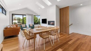A modern living room with a wooden dining table, wicker chairs, a sofa, and a TV mounted on the wall. - Luxury Riverside Escape with Fire Pit, Pizza Oven, River Views (Ocean Grove)