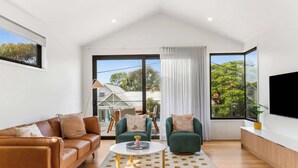 A modern living room with a brown leather sofa, two green armchairs, a white coffee table, and a large TV on the wall. - Luxury Riverside Escape with Fire Pit, Pizza Oven, River Views (Ocean Grove)