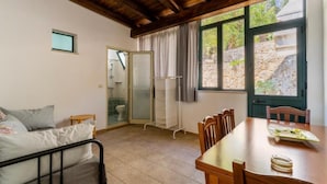A room with a bed, a table, and chairs, a shower area, and a view of a stone wall and greenery outside.