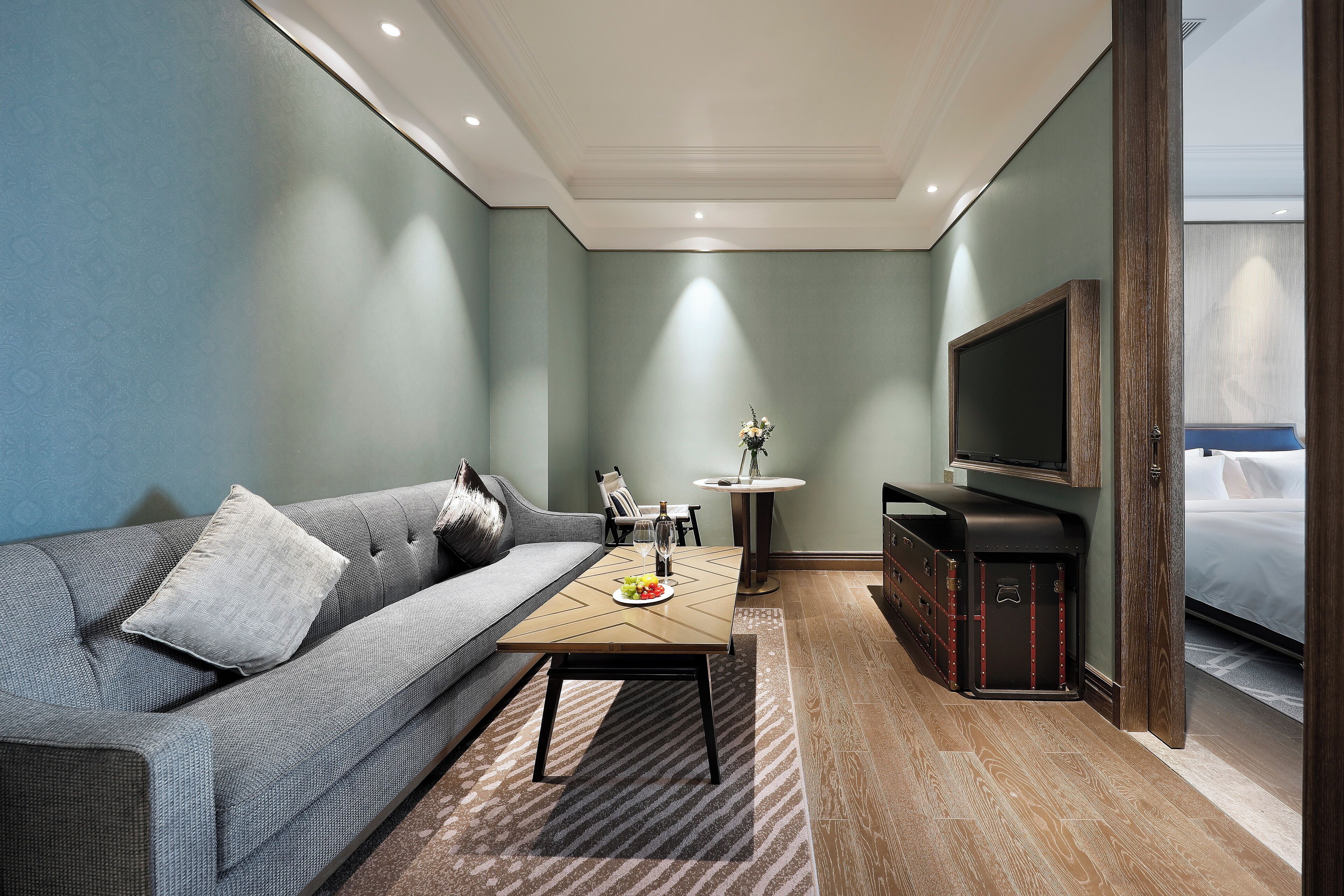 Una habitación de hotel moderna con un sofá gris, una mesa de centro de madera, un televisor y una cama.
