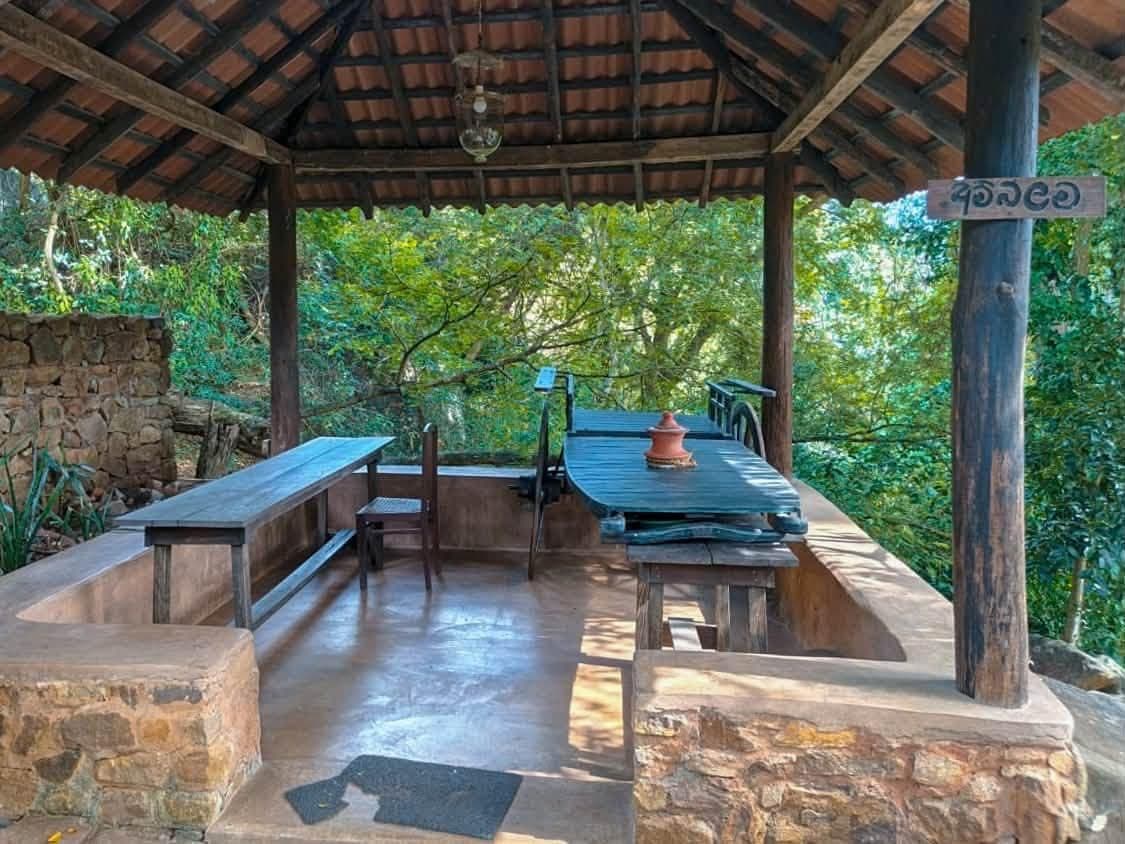 An outdoor covered seating area with stone benches and tables, surrounded by greenery.
