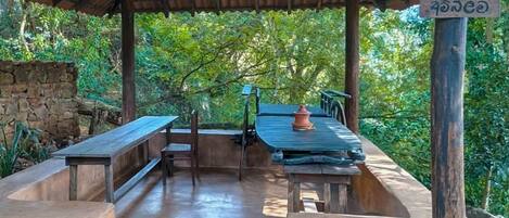An outdoor covered seating area with stone benches and tables, surrounded by greenery.