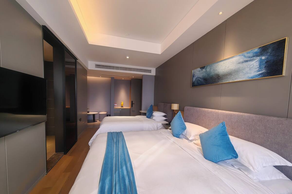A modern hotel room with a large bed, a flat-screen TV, a painting on the wall, and a bathroom visible in the background.