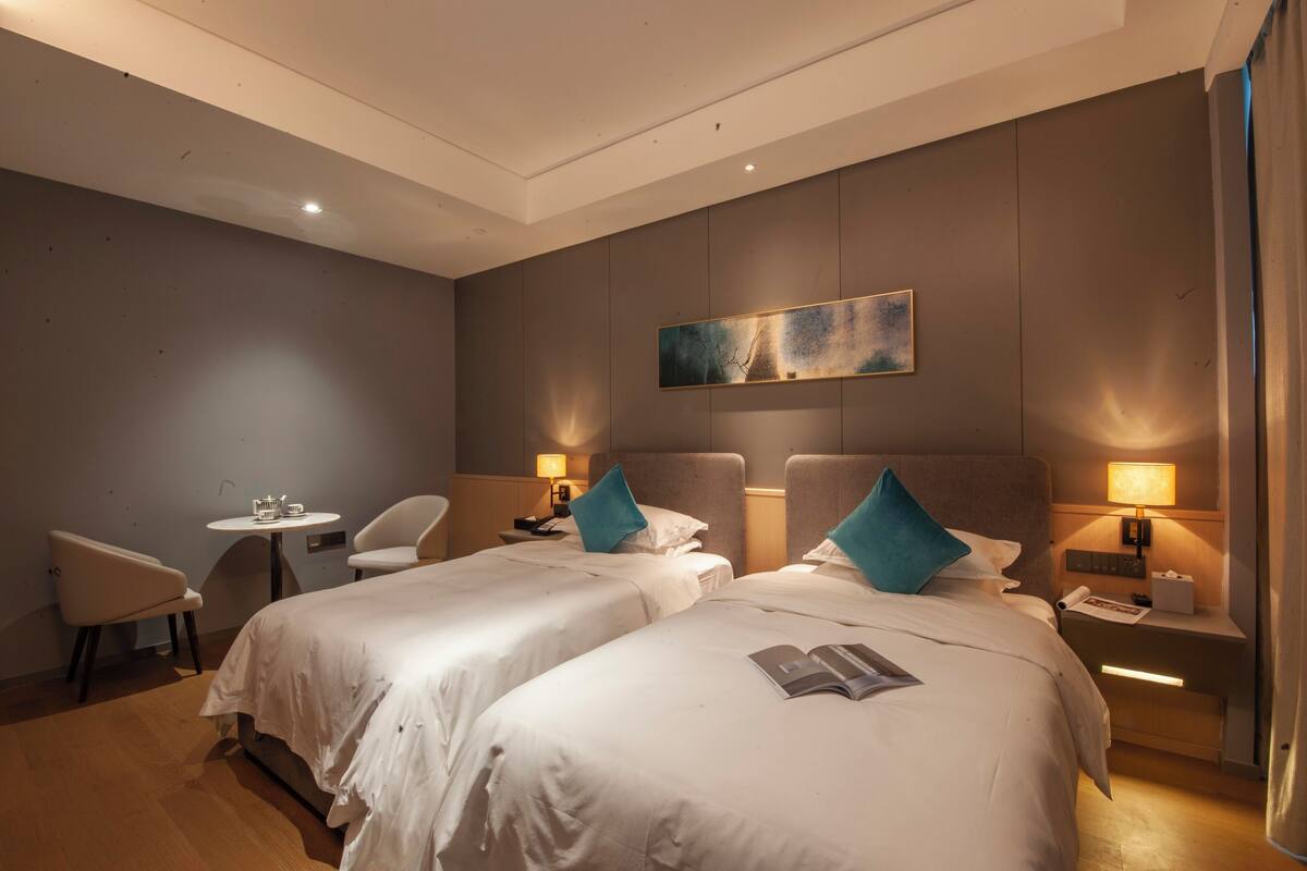 A hotel room with two beds, a small table, a chair, and a painting on the wall.