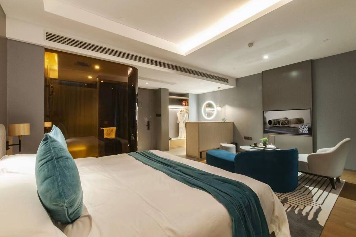 A modern hotel room with a large bed, a seating area with a blue sofa and armchairs, a small table, and a TV mounted on the wall.